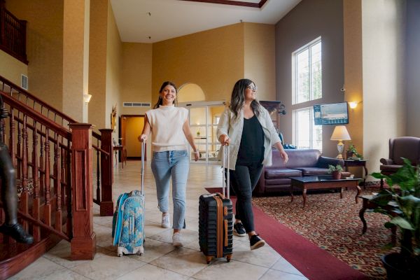 Two people with suitcases walking through a hotel lobby with seating, plants, and natural light from large windows.