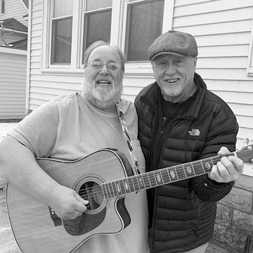 Two men smiling, with one holding a guitar and the other wearing a cap, standing in front of a house.